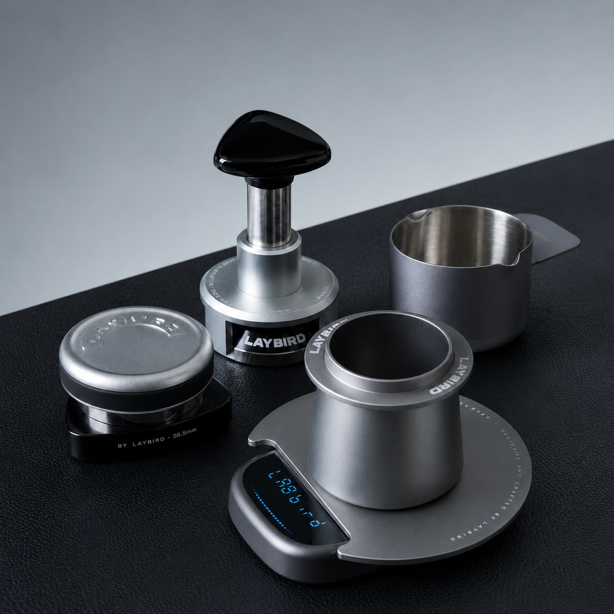 A coffee preparation set on a dark surface, including a digital scale with a metal portafilter holder, a tamper with a black handle, a small metal cup, and a round metal container. The products are shown cleanly with no people or real environment context beyond the tabletop and backdrop.